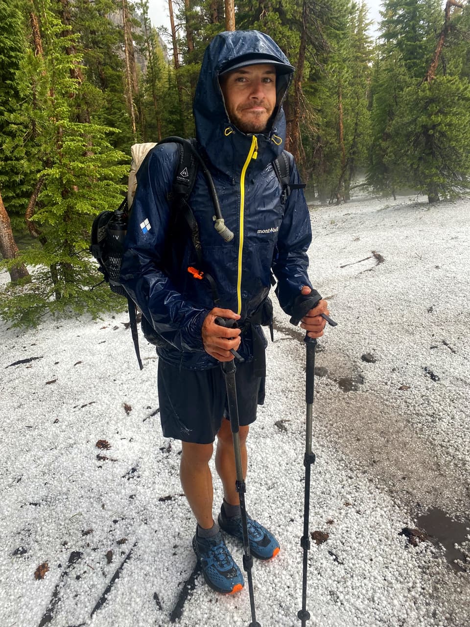 Atlas soaked in a navy Montbell rain jacket with trekking poles, standing in a field of hail