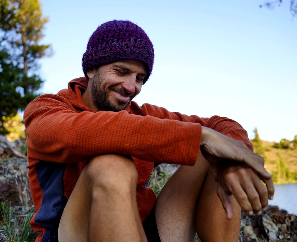 Atlas smiling in a purple crocheted beanie and orange hoodie by a lake at golden hour