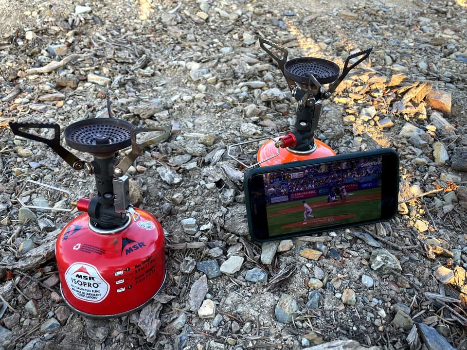 two MSR PocketRocket stoves on IsoPro canisters on gravel, a phone between them playing a ball game