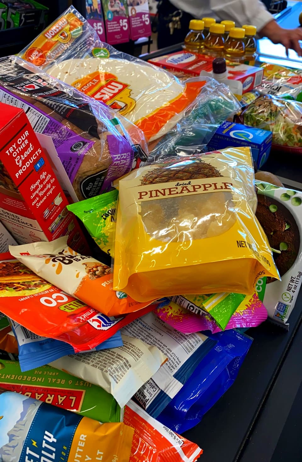 flour tortillas, dried pineapple, Larabars and other bars piled on a grocery checkout belt