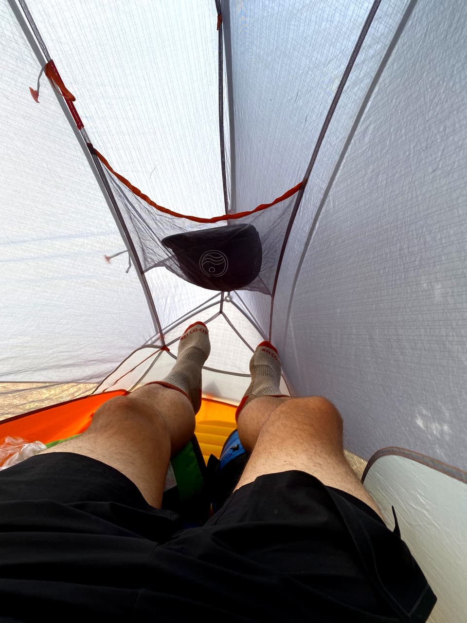 looking down the inside of a one-person tent, legs on an orange sleeping pad, a cap in the mesh gear loft